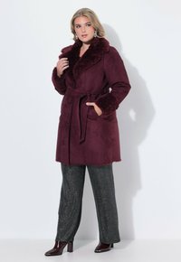 Burgunderfärgad fuskmockakappa med en fluffig krage och manschetter, bälte i midjan och två knappar. Tillsammans med glittrande grå byxor och blanka svarta stövlar.