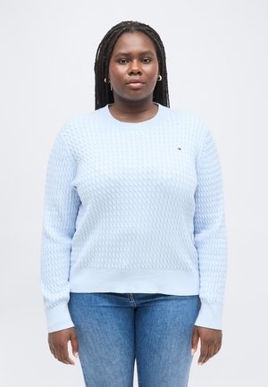 Kvinde med langt fletning hår iført gyldne øreringe, en lyseblå tekstureret sweater og blå jeans, stående mod en ensfarvet hvid baggrund.