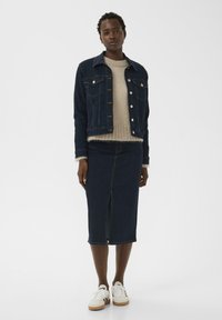 Dunkle Jeansjacke mit Vordertaschen, ein hellbeiger Strickpullover und ein passender Jeans-Bleistiftrock mit Schlitz vorne, kombiniert mit weißen Turnschuhen.