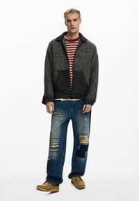 Männerjacke aus strukturiertem, dunklem Material mit Kragen über einem gestreiften rot-weißen Hemd. Blaue, ausgewaschene Jeans mit verschiedenen Applikationsdetails. Braune Schuhe.