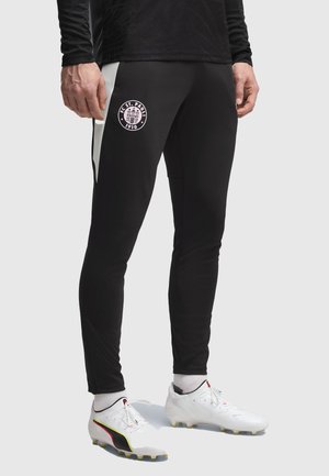 Personne portant un pantalon de sport noir FC St. Pauli 1910 avec des bandes blanches sur les côtés et des crampons blancs avec des chaussettes blanches sur un fond uni.