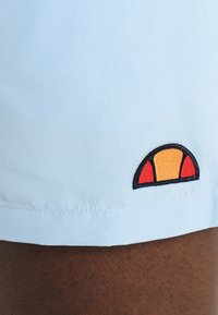 Lyseblå badebukser med en glatt tekstur, som har en brodert logo i oransje, gul og rød på kanten.
