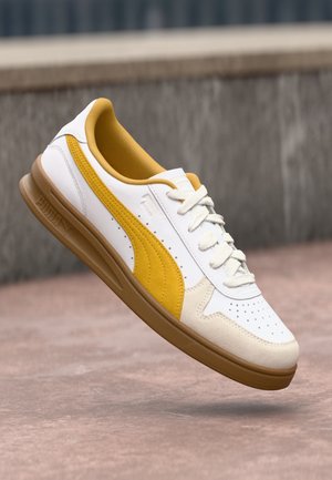 White sneaker with yellow accents and perforated detailing, featuring a textured sole and lace-up design. Logo embossed on the side.