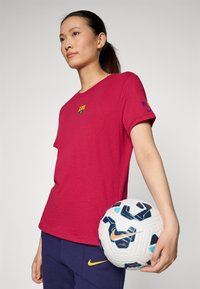 Burgunderfarget t-skjorte med FC Barcelona-logo på brystet, kombinert med marineblå bukser med en gul Nike-swoosh; holder et hvitt og blåmønstret fotball.