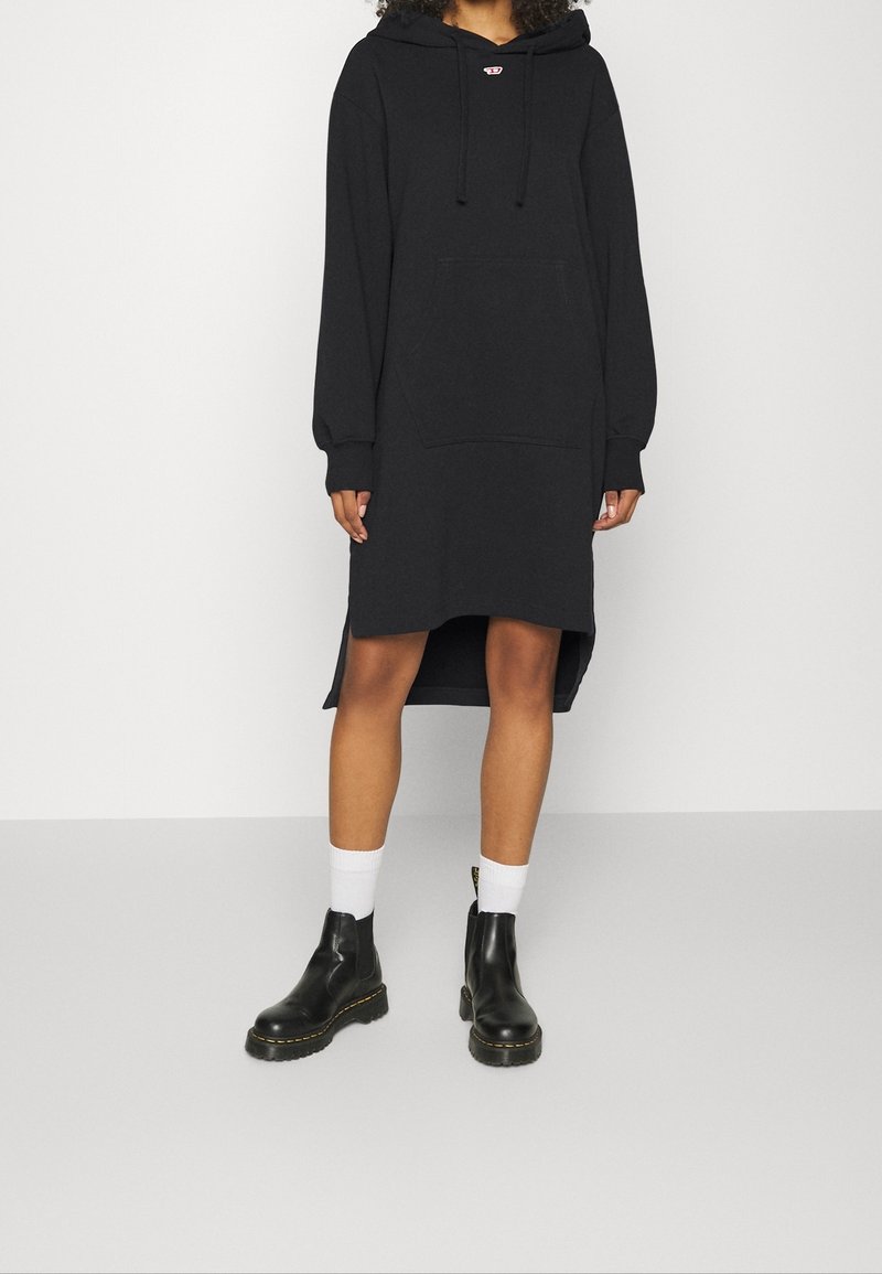 Svart huvtröja klänning med långa ärmar, framficka och sidoklyftor; bärs med svarta ankelkängor och vita strumpor.