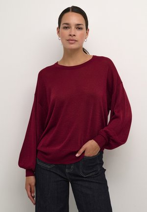 Burgundská oversized pletená svetr s dlouhými nadýchanými rukávy, ribbovanými manžetami a kulatým výstřihem, v kombinaci s tmavými džíny.