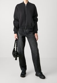 Veste bomber noire zippée avec des poignets côtelés, assortie à un jean en denim foncé à coupe ample et des baskets noires. Portant un petit sac à main noir.