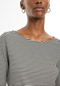 Zwart-wit gestreepte ribbeltop met een wijde halslijn en lange mouwen, met een gladde textuur en subtiele glans. Gouden ringoorbellen zichtbaar.