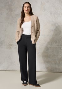 Cardigan en tricot beige avec trois boutons, associé à un pantalon noir taille haute et des baskets noires avec des lacets blancs et des semelles brunes.