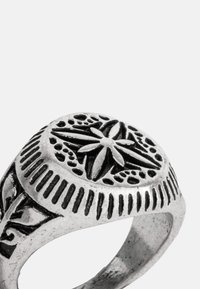 Anel prateado com um design circular, apresentando padrões florais intrincados e acentos texturizados em uma faixa larga. O acabamento mate realça os detalhes.