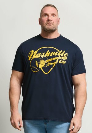 Muskelbepackter Mann trägt ein dunkelblaues T-Shirt mit gelbem Schriftzug "Nashville Music City" und Gitarrengrafik, steht vor einem schlichten grauen Hintergrund.
