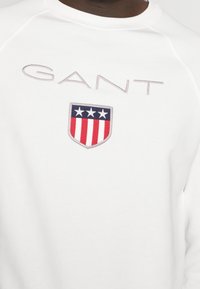 Sudadera blanca con el logo de GANT, que presenta un diseño de escudo con rayas rojas y tres estrellas azules. Tejido suave y texturizado.