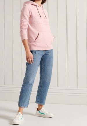 Persoon draagt een roze hoodie, blauwe cropped jeans en witte sneakers met turquoise accenten, staand tegen een witte paneelmuur.
