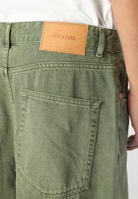 Calças verde-azeitona com um tecido texturizado. Apresenta uma etiqueta de couro rotulada "ONLY & SONS" na cintura e um design no bolso traseiro.