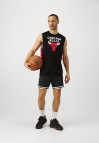 Melna bezsvārku krekls ar tekstu "CHICAGO BULLS" un bulla logo, apvienots ar melniem šortiem, kuriem ir baltas sānu svītras; tur rokās basketbola bumbu.