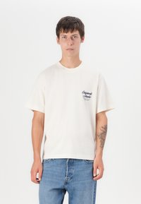 T-shirt en coton crème avec un col rond, des manches courtes et un petit motif bleu sur la poitrine gauche. Porté avec un jean en denim bleu.