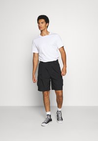 Pantalones cortos de carga negros con bolsillos laterales, combinados con una camiseta blanca básica y zapatillas negras de caña alta, contra un fondo neutro.