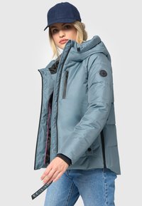 Lichtblauwe pufferjack met een capuchon, voorzien van een zwarte rits, zijzakken en een logopatch op de mouw. Draag het met denim jeans.