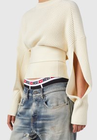 Suéter de punto acanalado crema con mangas largas, diseño corto y aberturas laterales. Combinado con jeans de denim claro desgastados y una cintura de marca.