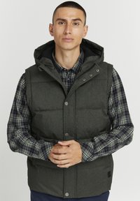 Padded vest in dark green fabric with a hood, featuring snap buttons and side pockets, worn over a plaid shirt with navy and green tones.
