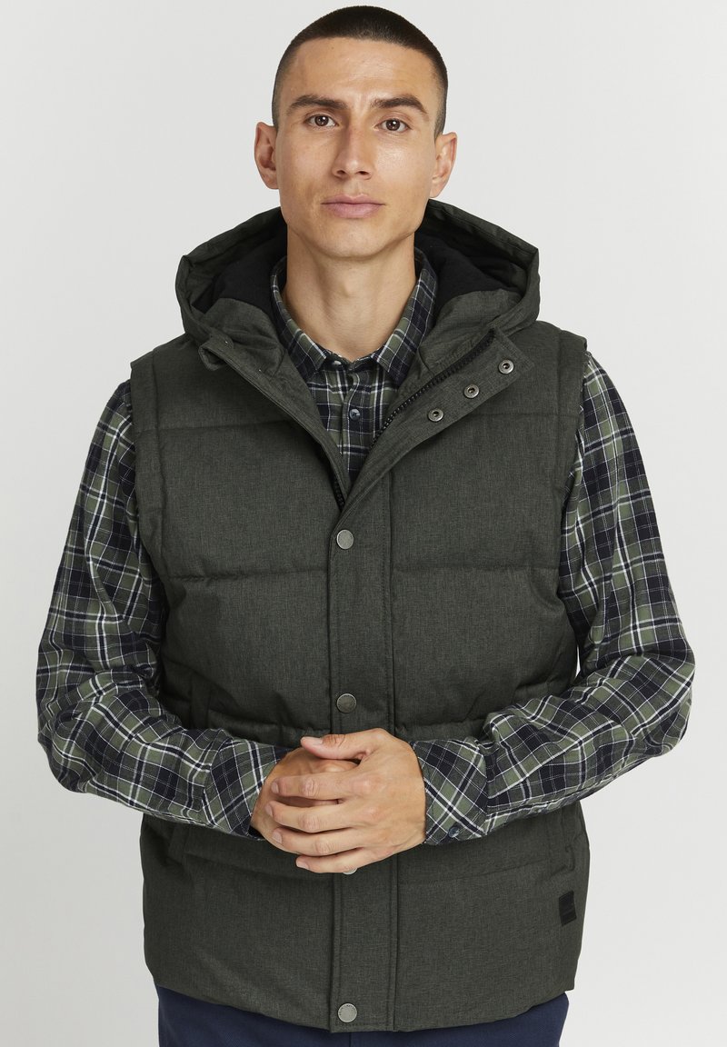 Padded vest in dark green fabric with a hood, featuring snap buttons and side pockets, worn over a plaid shirt with navy and green tones.
