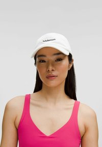 Biała czapka baseballowa z haftem "lululemon" w kolorze czarnym. Noszona z różową bezrękawnikową bluzką. Gładki materiał, strukturalny fason, minimalistyczny styl.