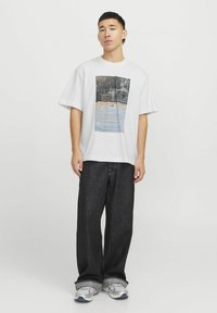 Camiseta blanca de manga corta con un gráfico rectangular de una cancha de baloncesto. Llevada con unos vaqueros de denim oscuros de corte suelto y zapatillas gris.