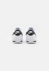 White Nike shoes featuring a black textured heel accent, rounded shape, and rubber outsole with a patterned tread. Classic logo on the heel.