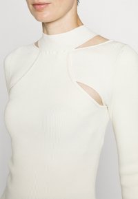 Maglia bianca a maniche lunghe con coste, caratterizzata da un collo alto, dettagli cut-out sulle spalle e una silhouette aderente. Tessuto morbido e materiale elasticizzato.