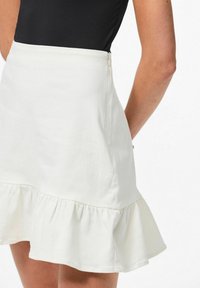 Baltas kokvilnas svārki ar izplestu ruffles apakšmalu, kas raksturojas ar gludu tekstūru un aizmugurēju rāvējslēdzēja aizdari. Nav redzamu rakstu vai akcentu.