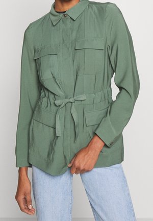Person iført en grøn langærmet jakke med lommer på bryst og talje og et bundet bælte i taljen, kombineret med lyseblå jeans.