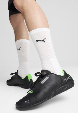 Czarne sportowe buty z zielonymi akcentami, z gładką, perforowaną powierzchnią i sznurowanym zapięciem. Noszone z białymi skarpetkami w prążki z logo.