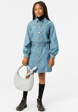 Denimklänning med långa ärmar, knappar fram och midja med dragsko. Kombinerad med en grå handväska och svarta knähöga strumpor.