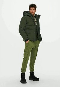 Giovane uomo che indossa una giacca imbottita verde scuro con cappuccio, un maglione a fantasia, pantaloni cargo oliva e stivali neri con lacci, in piedi con le mani nelle tasche.