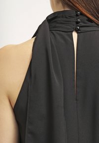 Vue arrière d'une femme portant une robe noire sans manches avec un col haut plissé maintenu par trois boutons noirs.