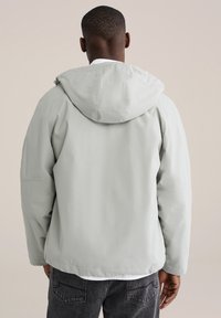 Lichtgrijs capuchonjack gemaakt van soepel, waterbestendig materiaal. Heeft een ontspannen pasvorm en geribbelde manchetten, met een minimalistisch ontwerp.