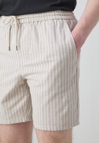 Short beige à fines rayures verticales, taille élastique avec cordon de serrage, porté avec la main droite dans la poche.