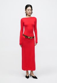 Rød strikket kjole med lange ærmer, tætsiddende silhuet, rund halsudskæring og et brunt bælte. Sorte spidse hæle fuldender outfittet.