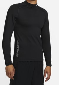 Mann in schwarzem Langarm-Kompressionsshirt mit dem Text "Nike Pro" an der Seite und passenden schwarzen Shorts, stehend vor einem weißen Hintergrund.