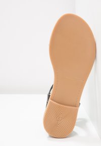 Sandaalin pohja, jossa on ruskea kumipohja, pintakuvioitu liukastumisen estämiseksi, merkitty "37," ja siinä on sileä, kaareva reuna sekä osittain näkyvä musta hihna.