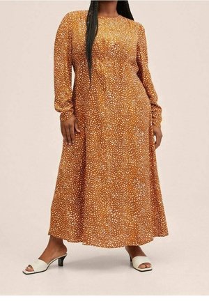 Femme portant une longue robe orange à taches blanches et des talons bas blancs ouverts sur un fond uni.