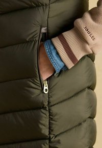 Ręka w kieszeni ciemnozielonej pikowanej kurtki z beżowym mankietem i widoczną dżinsową koszulą pod spodem.