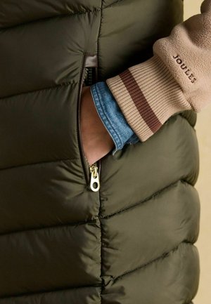 Hand in der Tasche einer dunkelgrünen Steppjacke mit beigem Ärmelbund und sichtbarem Jeanshemd darunter.