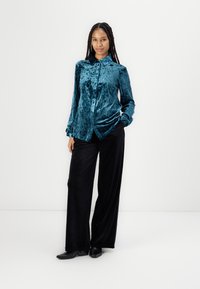 Teal velvet button-up shirt with a collared neck, long sleeves, and black wide-leg trousers. Smooth texture with shiny finish.