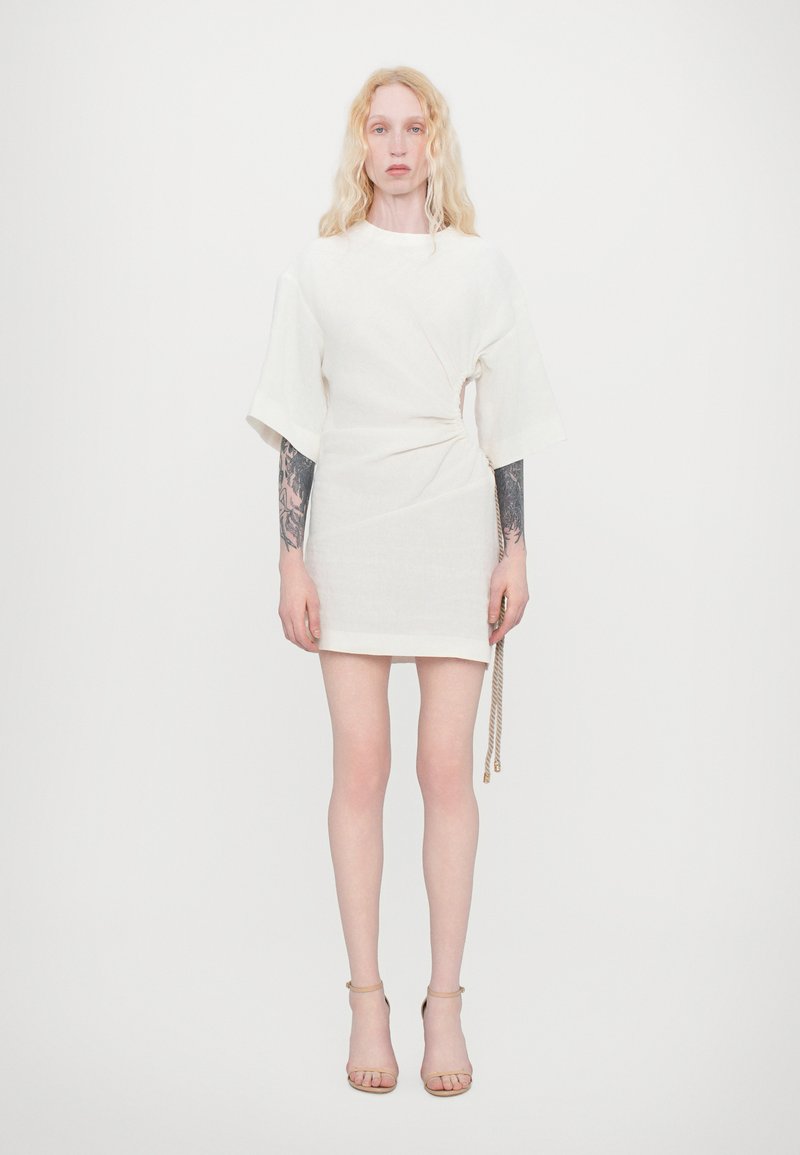 White short dress with a gathered design, wide sleeves, and a side loop detail. Smooth fabric with a subtle texture. Footwear: tan strappy heels.