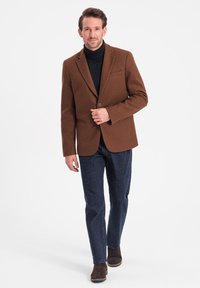 Brun ullblazer med enkel knappstängning, slagkragar och bröstficka, bärs över en svart polotröja och matchas med mörk denimjeans.