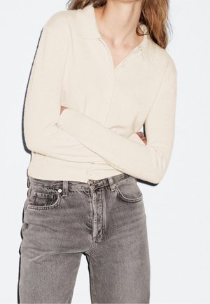 Femme portant un cardigan crème à manches longues boutonné et un jean gris délavé taille haute, debout devant un fond blanc.