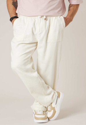 Homme portant un pantalon blanc ample, des baskets beige et blanc, et une chemise rose clair, les mains dans les poches, debout jambes croisées.