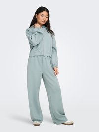 Lichtblauwe capuchontrui met rits en elastische boorden, gecombineerd met loszittende bijpassende broek. Gladde stof met een losse pasvorm.