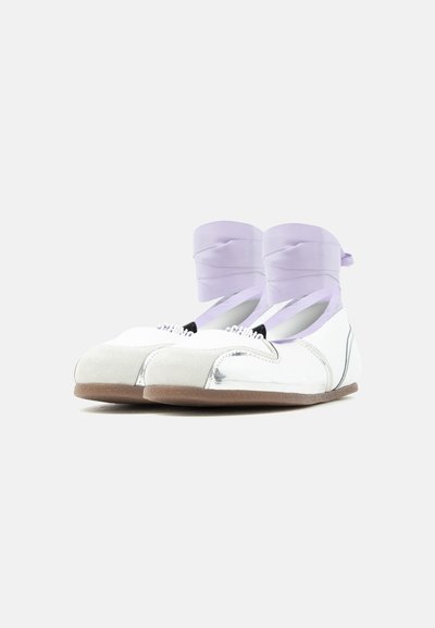 Ballerines blanches avec bouts en suède, semelles marron et rubans lavande noués autour des chevilles, sur un fond uni.